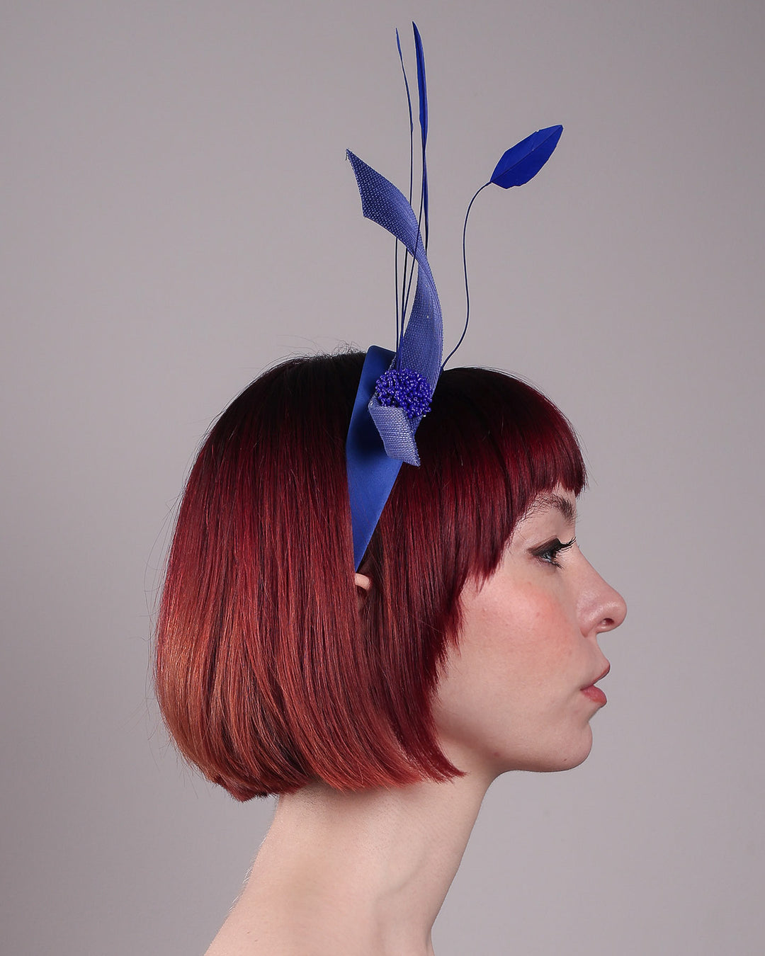 The model showcases the right side close up of the Motmot headband fascinator crafted in a rich cobalt blue. This bold headband features a spray of sculpted feathers with hand-curved edges, accented with a swirl of buntal ribbon tied together with a beaded centre. Designed by Merve Bayindir.