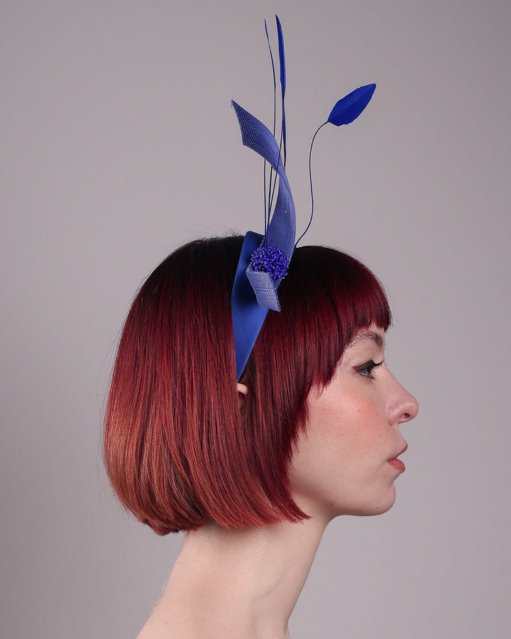 The model showcases the right side close up of the Motmot headband fascinator crafted in a rich cobalt blue. This bold headband features a spray of sculpted feathers with hand-curved edges, accented with a swirl of buntal ribbon tied together with a beaded centre. Designed by Merve Bayindir.