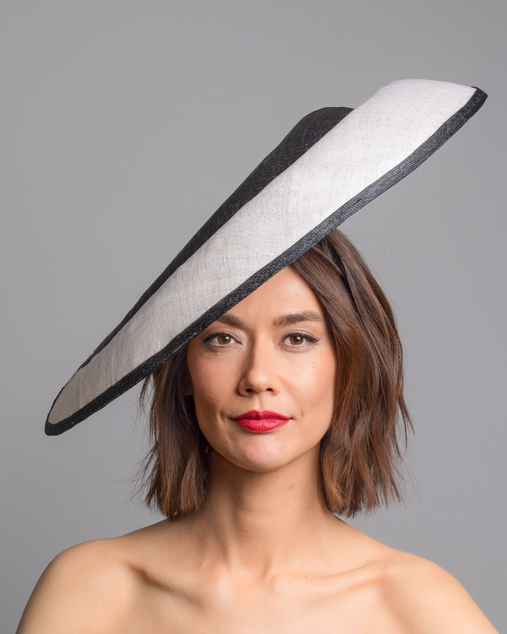 The model is showcasing the front side close up of the wide black and white hat with an oversized flat brim, featuring a white woven top edged in black trim and finished with a black floral accent and slim headband for secure wear. Designed and made by Merve Bayindir MIllinery.
