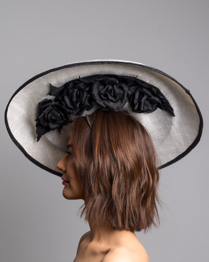 The model is showcasing the left side close up of the wide black and white hat with an oversized flat brim, featuring a white woven top edged in black trim and finished with a black floral accent and slim headband for secure wear. Designed and made by Merve Bayindir MIllinery.
