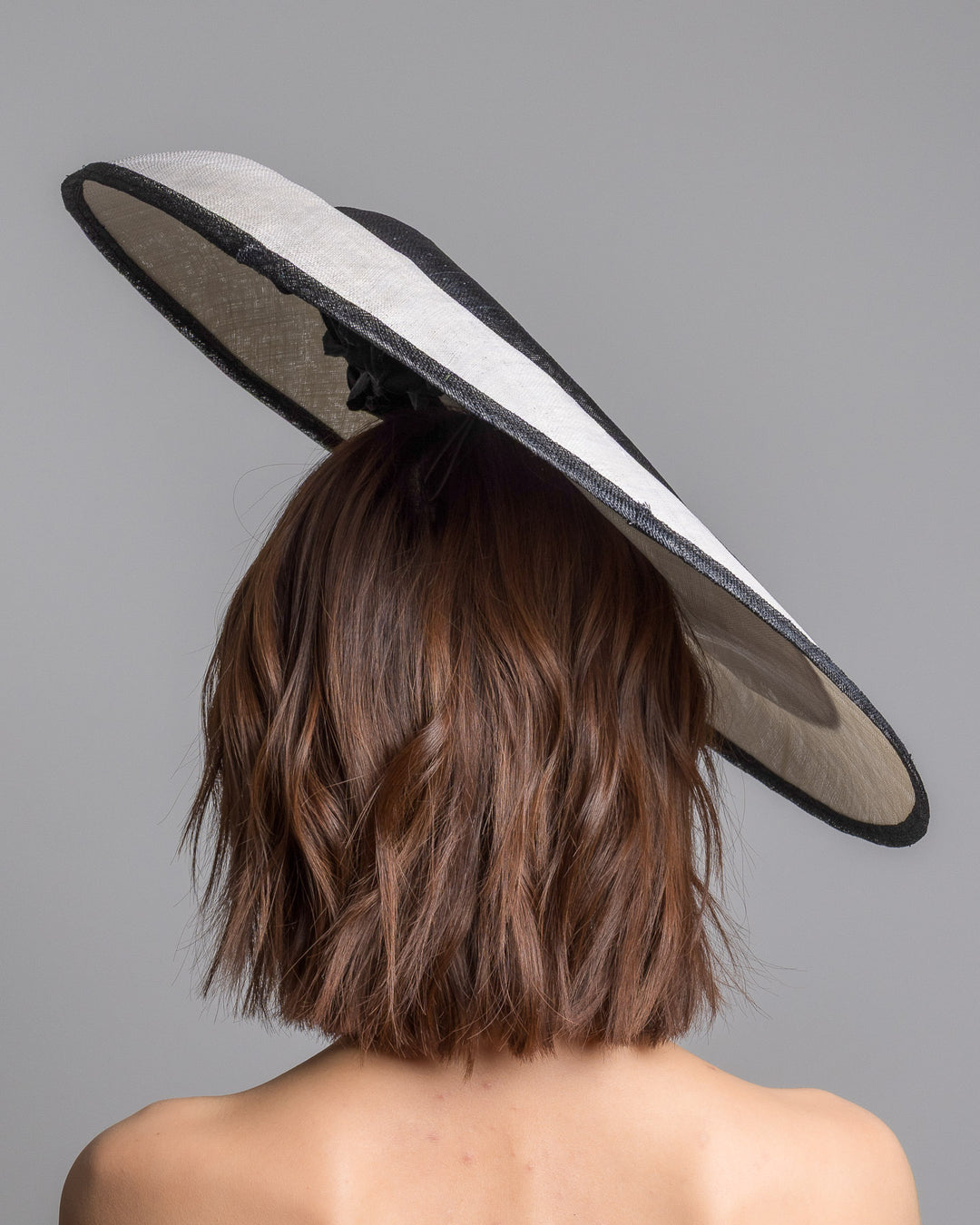 The model is showcasing the back side close up of the wide black and white hat with an oversized flat brim, featuring a white woven top edged in black trim and finished with a black floral accent and slim headband for secure wear. Designed and made by Merve Bayindir MIllinery.
