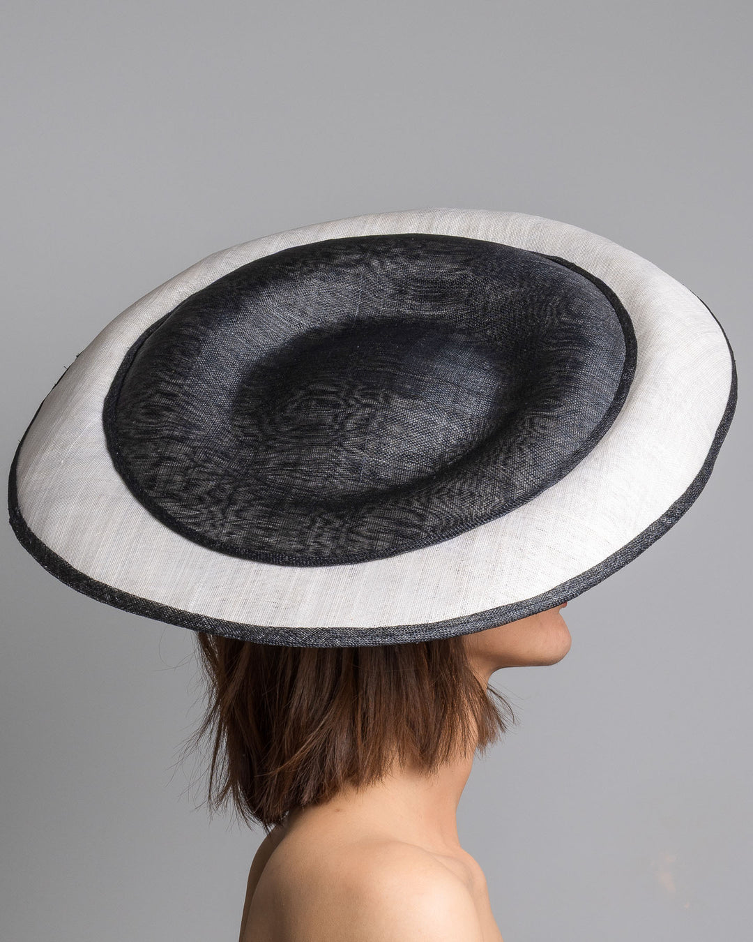 The model is showcasing the right side close up of the wide black and white hat with an oversized flat brim, featuring a white woven top edged in black trim and finished with a black floral accent and slim headband for secure wear. Designed and made by Merve Bayindir MIllinery.
