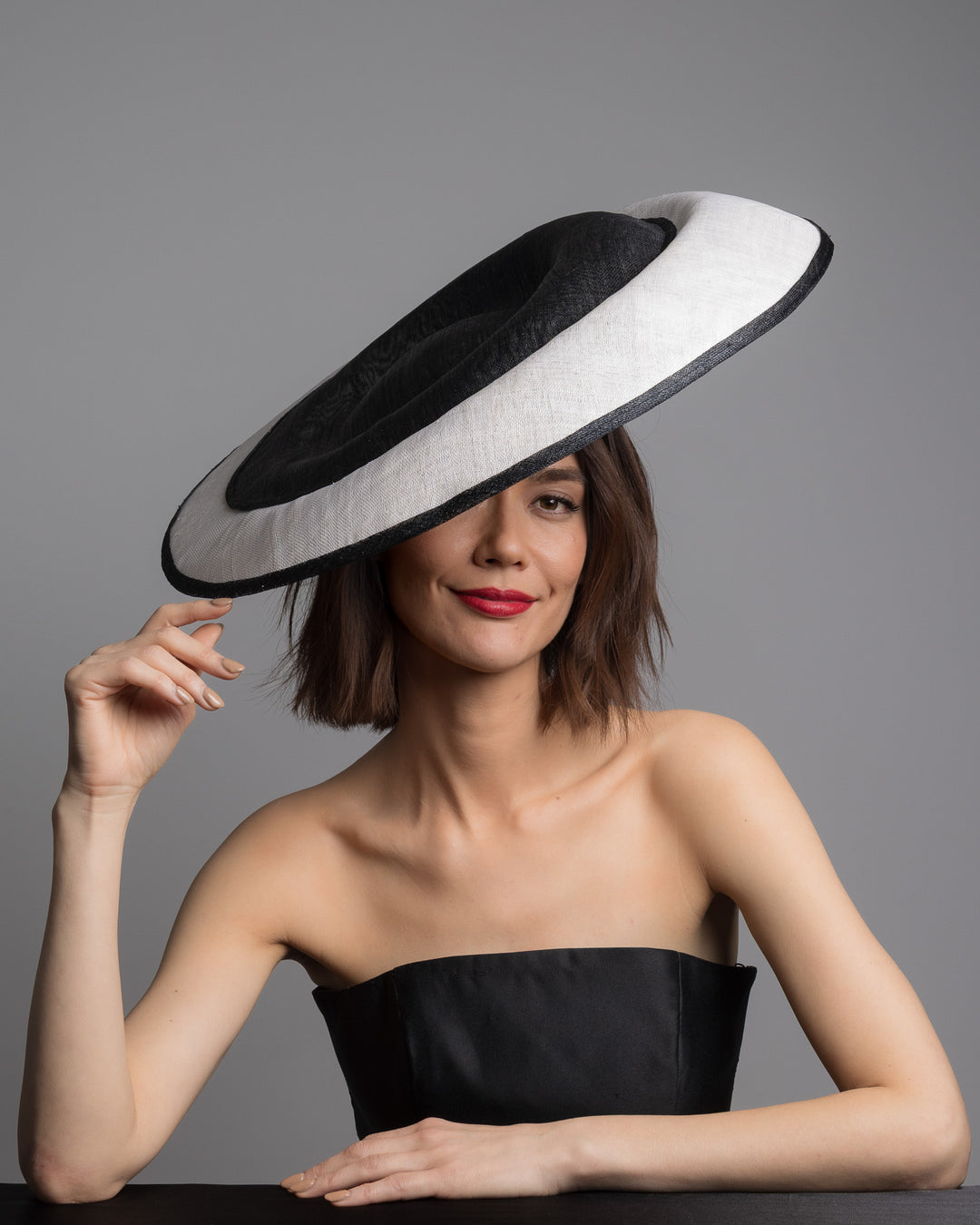 The female model is posing, wearing 
Noctel, the wide black and white hat with an oversized flat brim, featuring a white woven top edged in black trim and finished with a black floral accent and slim headband for secure wear. Designed and made by Merve Bayindir MIllinery.
