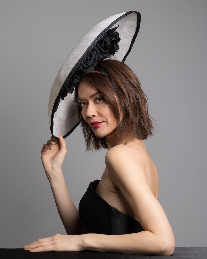 The female model is posing, wearing 
Noctel, the wide black and white hat with an oversized flat brim, featuring a white woven top edged in black trim and finished with a black floral accent and slim headband for secure wear. Designed and made by Merve Bayindir MIllinery.
