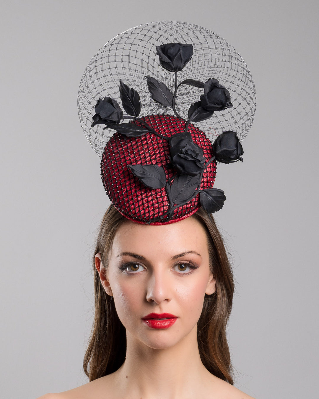 The model is showcasing the front side close up of the bold fascinator composed of a structured circular base in deep red, overlaid with black netting that creates a texture. From the base an  arrangement of matte black roses and elongated leaves, climbing upward in a graceful, vine-like composition.Behind the florals, a sheer circular veil of fine netting forms a delicate halo. Designed and made by Merve Bayindir Millinery.
