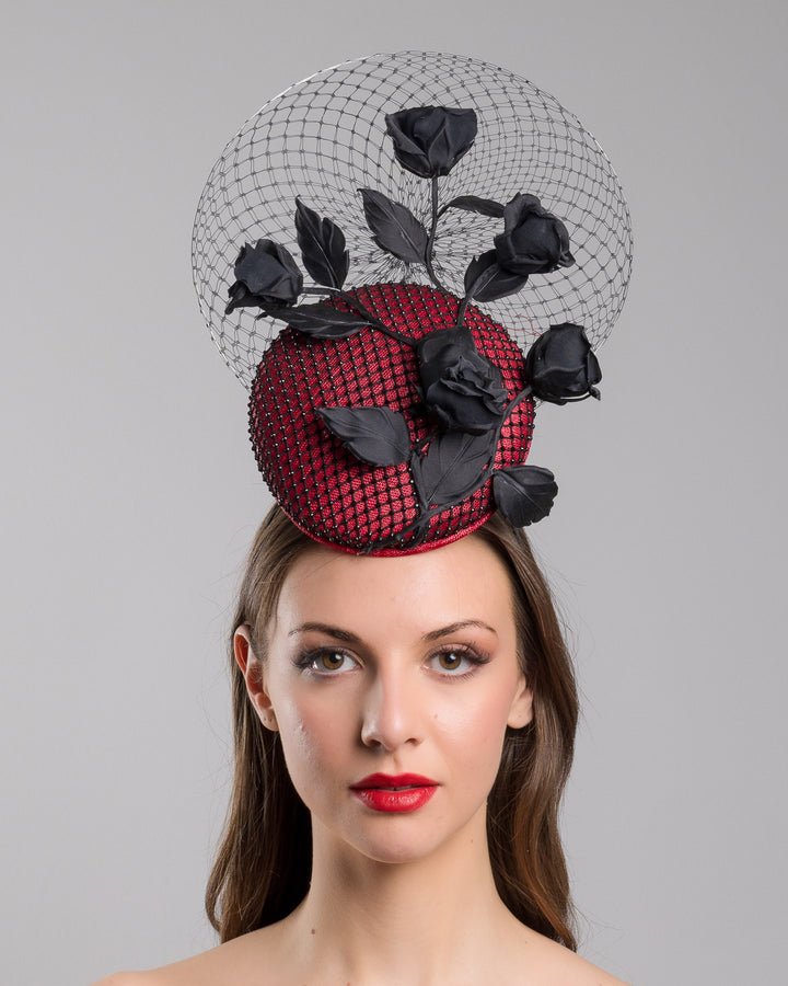 The model is showcasing the front side close up of the bold fascinator composed of a structured circular base in deep red, overlaid with black netting that creates a texture. From the base an  arrangement of matte black roses and elongated leaves, climbing upward in a graceful, vine-like composition.Behind the florals, a sheer circular veil of fine netting forms a delicate halo. Designed and made by Merve Bayindir Millinery.
