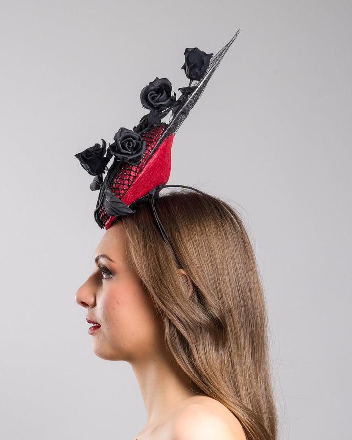 The model is showcasing the left side close up of the bold fascinator composed of a structured circular base in deep red, overlaid with black netting that creates a texture. From the base an  arrangement of matte black roses and elongated leaves, climbing upward in a graceful, vine-like composition.Behind the florals, a sheer circular veil of fine netting forms a delicate halo. Designed and made by Merve Bayindir Millinery.

