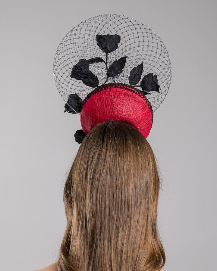 The model is showcasing the back side close up of the bold fascinator composed of a structured circular base in deep red, overlaid with black netting that creates a texture. From the base an  arrangement of matte black roses and elongated leaves, climbing upward in a graceful, vine-like composition.Behind the florals, a sheer circular veil of fine netting forms a delicate halo. Designed and made by Merve Bayindir Millinery.
