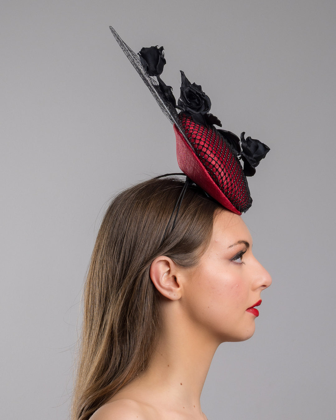 The model is showcasing the right side close up of the bold fascinator composed of a structured circular base in deep red, overlaid with black netting that creates a texture. From the base an  arrangement of matte black roses and elongated leaves, climbing upward in a graceful, vine-like composition.Behind the florals, a sheer circular veil of fine netting forms a delicate halo. Designed and made by Merve Bayindir Millinery.
