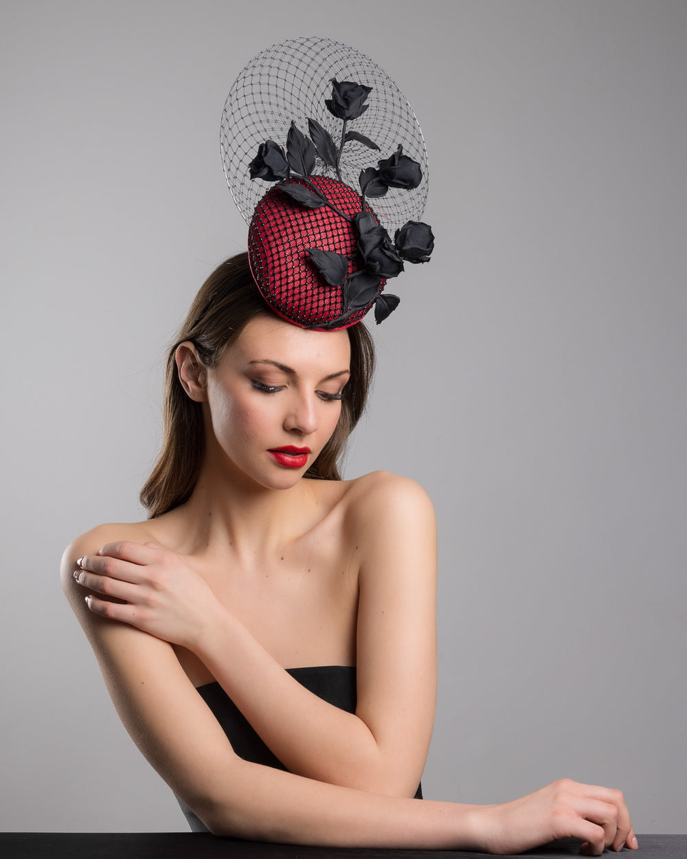 The female model is posing, wearing
Nocte, the bold fascinator composed of a structured circular base in deep red, overlaid with black netting that creates a texture. From the base an  arrangement of matte black roses and elongated leaves, climbing upward in a graceful, vine-like composition.Behind the florals, a sheer circular veil of fine netting forms a delicate halo. Designed and made by Merve Bayindir Millinery.
