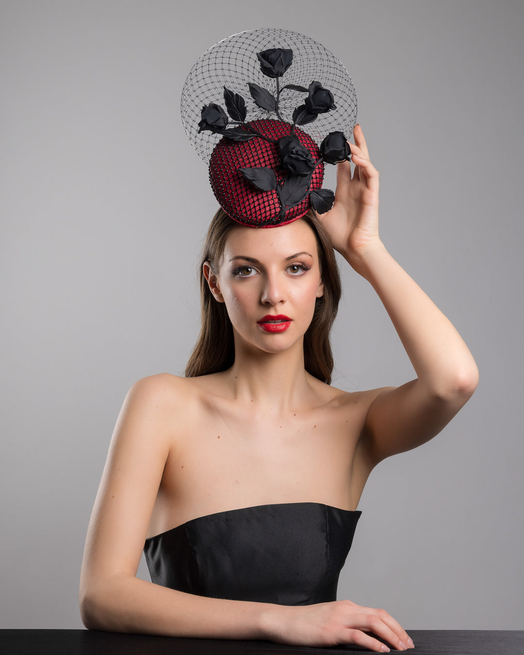 The female model is posing, wearing
Nocte, the bold fascinator composed of a structured circular base in deep red, overlaid with black netting that creates a texture. From the base an  arrangement of matte black roses and elongated leaves, climbing upward in a graceful, vine-like composition.Behind the florals, a sheer circular veil of fine netting forms a delicate halo. Designed and made by Merve Bayindir Millinery.
