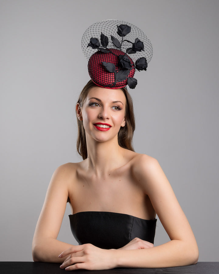 The female model is posing, wearing
Nocte, the bold fascinator composed of a structured circular base in deep red, overlaid with black netting that creates a texture. From the base an  arrangement of matte black roses and elongated leaves, climbing upward in a graceful, vine-like composition.Behind the florals, a sheer circular veil of fine netting forms a delicate halo. Designed and made by Merve Bayindir Millinery.
