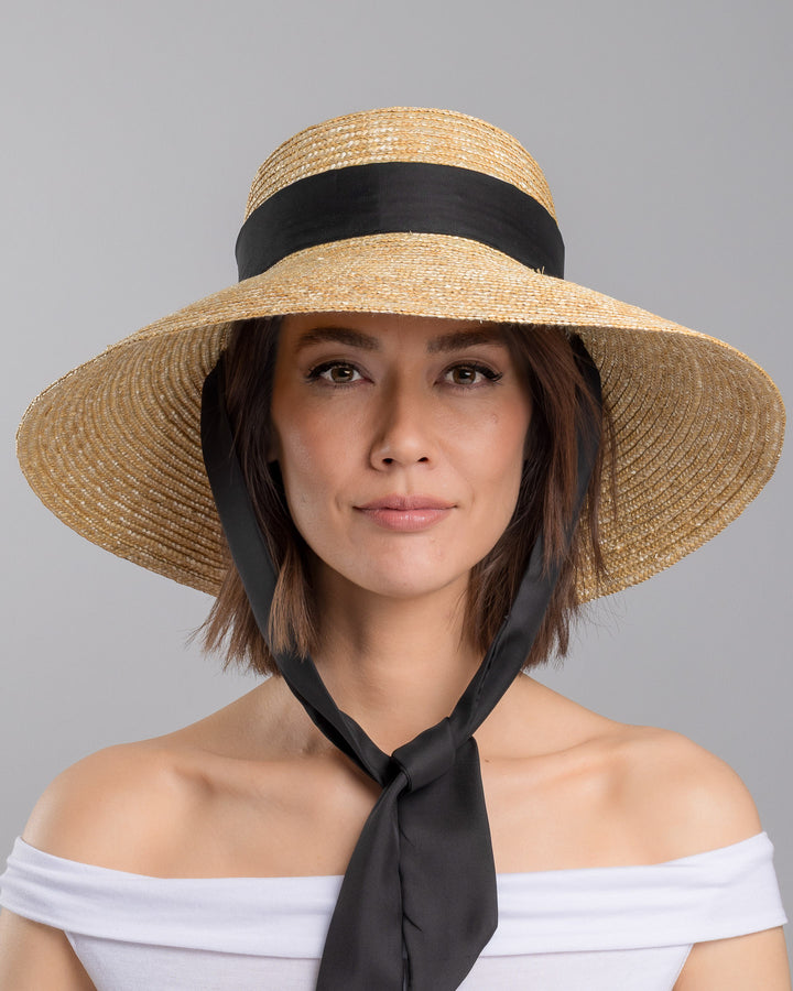 The model is showcasing the front side close up of the natural woven straw wide brim boater hat with a wide brim, wrapped in a black ribbon and finished with an oversized black satin bow at the side. Designed and made by Merve Bayindir Millinery.

