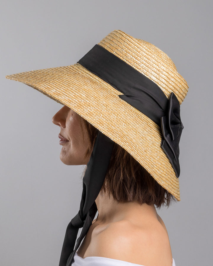 The model is showcasing the left side close up of the natural woven straw wide brim boater hat with a wide brim, wrapped in a black ribbon and finished with an oversized black satin bow at the side. Designed and made by Merve Bayindir Millinery.

