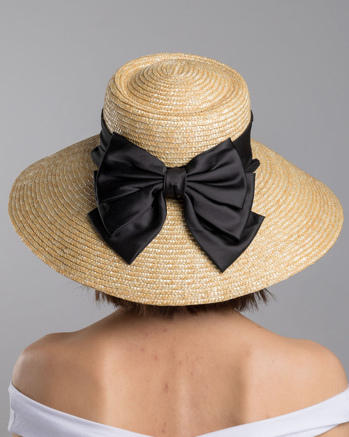 The model is showcasing the back side close up of the natural woven straw wide brim boater hat with a wide brim, wrapped in a black ribbon and finished with an oversized black satin bow at the side. Designed and made by Merve Bayindir Millinery.
