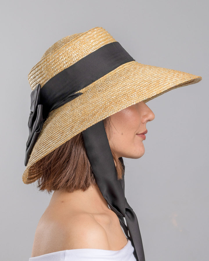The model is showcasing the right side close up of the natural woven straw wide brim boater hat with a wide brim, wrapped in a black ribbon and finished with an oversized black satin bow at the side. Designed and made by Merve Bayindir Millinery.
