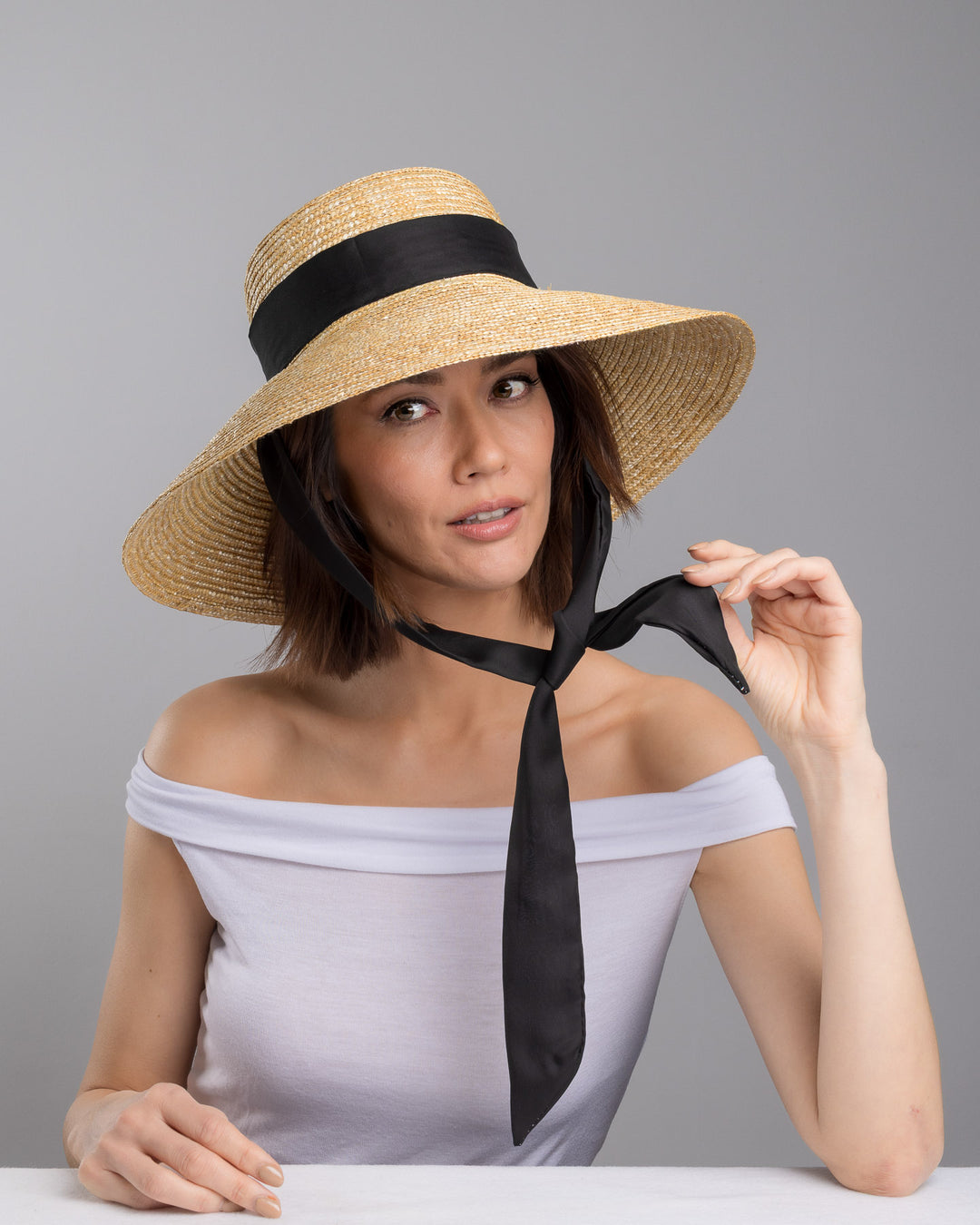 The  female model is posing, wearing Noen, the natural woven straw wide brim boater hat with a wide brim, wrapped in a black ribbon and finished with an oversized black satin bow at the side. Designed and made by Merve Bayindir Millinery.