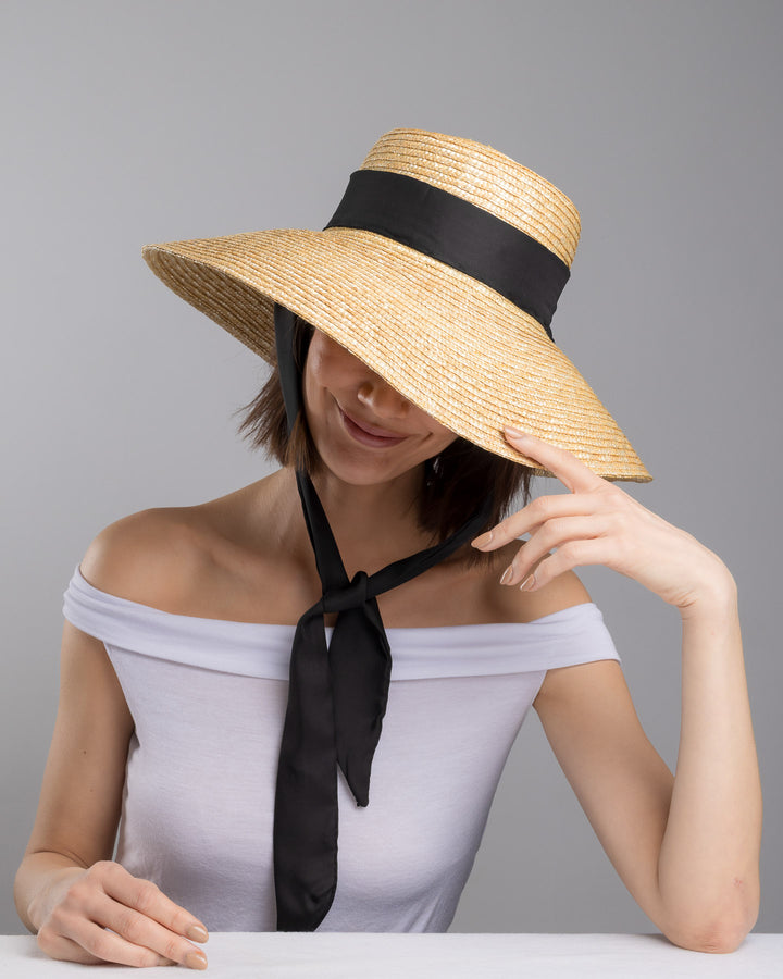 The  female model is posing, wearing Noen, the natural woven straw wide brim boater hat with a wide brim, wrapped in a black ribbon and finished with an oversized black satin bow at the side. Designed and made by Merve Bayindir Millinery.

