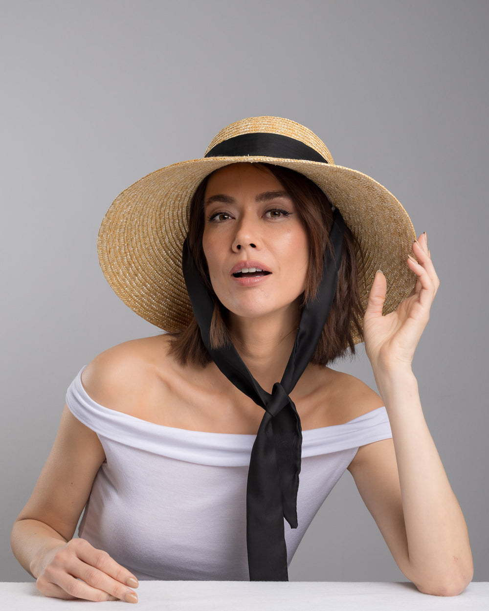 The  female model is posing, wearing Noen, the natural woven straw wide brim boater hat with a wide brim, wrapped in a black ribbon and finished with an oversized black satin bow at the side. Designed and made by Merve Bayindir Millinery.