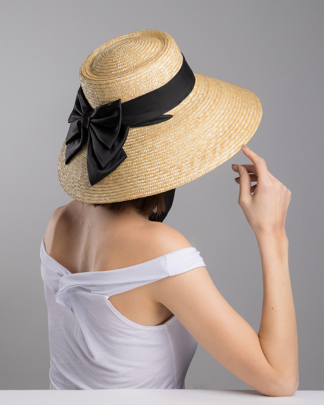 The  female model is posing, wearing Noen, the natural woven straw wide brim boater hat with a wide brim, wrapped in a black ribbon and finished with an oversized black satin bow at the side. Designed and made by Merve Bayindir Millinery.