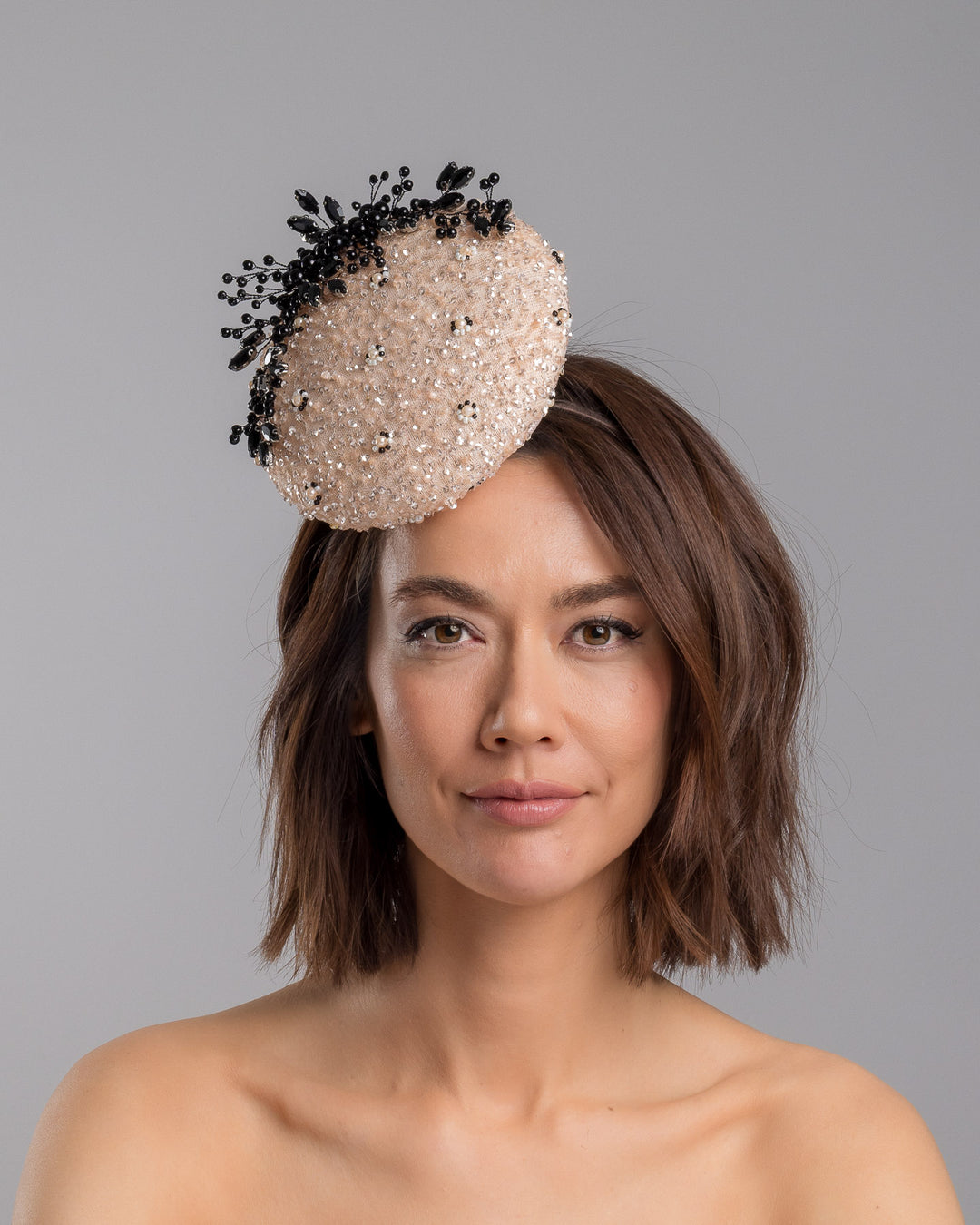 The model is showcasing the front side close up of the blush sinamay fascinator with a rounded high-back silhouette, intricately embellished with clear crystals and pearl accents, and finished with a dramatic black beaded floral spray for elegant contrast. Designed and made by Merve Bayindir Millinery.