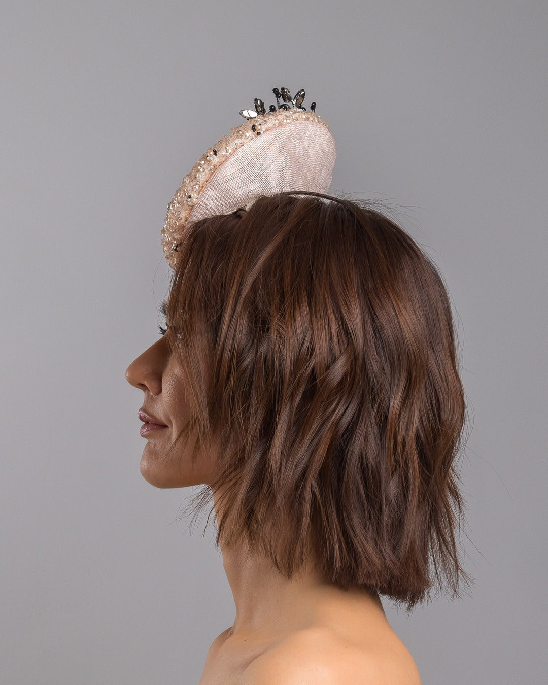 The model is showcasing the left side close up of the blush sinamay fascinator with a rounded high-back silhouette, intricately embellished with clear crystals and pearl accents, and finished with a dramatic black beaded floral spray for elegant contrast. Designed and made by Merve Bayindir Millinery.