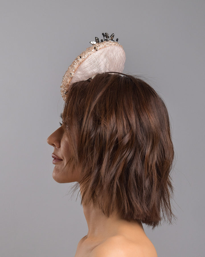 The model is showcasing the left side close up of the blush sinamay fascinator with a rounded high-back silhouette, intricately embellished with clear crystals and pearl accents, and finished with a dramatic black beaded floral spray for elegant contrast. Designed and made by Merve Bayindir Millinery.