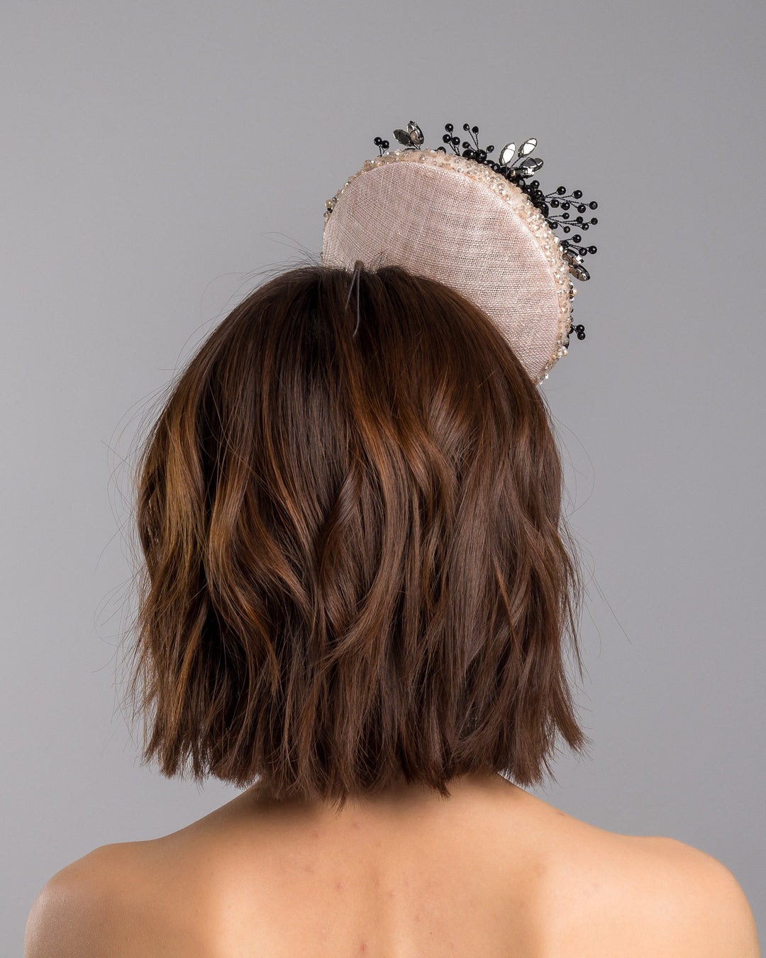 The model is showcasing the back side close up of the blush sinamay fascinator with a rounded high-back silhouette, intricately embellished with clear crystals and pearl accents, and finished with a dramatic black beaded floral spray for elegant contrast. Designed and made by Merve Bayindir Millinery.