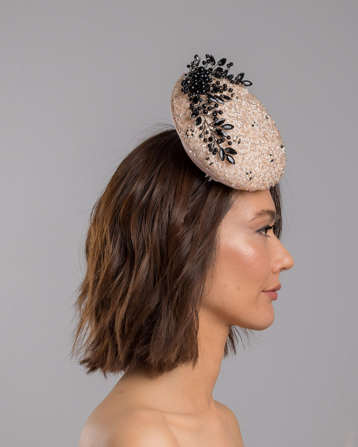 The model is showcasing the right side close up of the blush sinamay fascinator with a rounded high-back silhouette, intricately embellished with clear crystals and pearl accents, and finished with a dramatic black beaded floral spray for elegant contrast. Designed and made by Merve Bayindir Millinery.