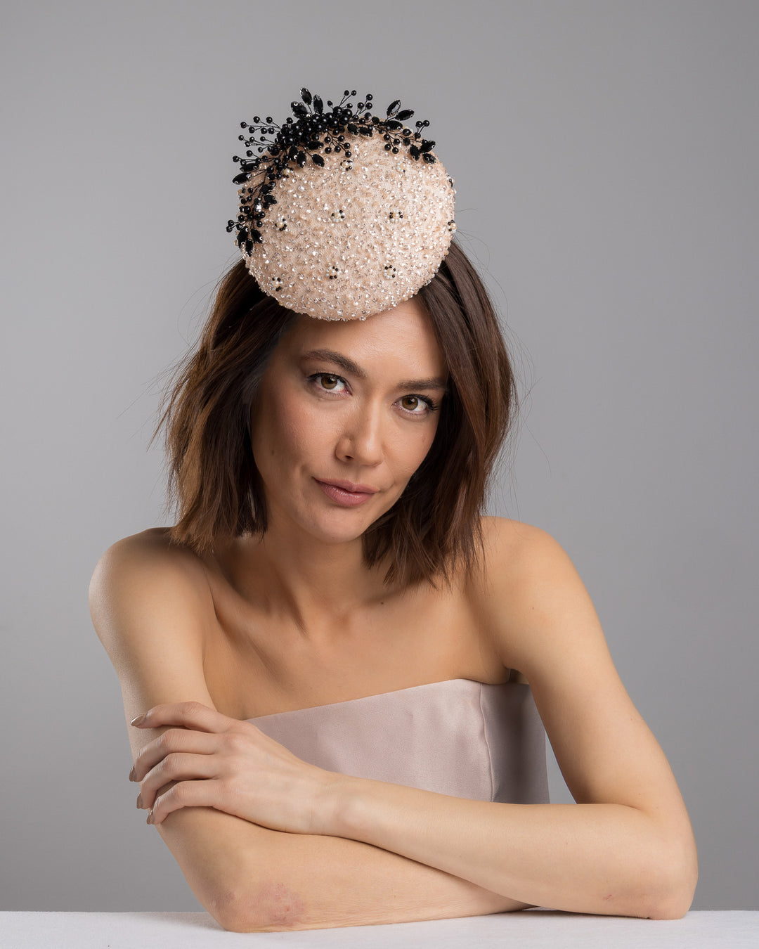 The female model is posing, wearing
Nuite, the blush sinamay fascinator with a rounded high-back silhouette, intricately embellished with clear crystals and pearl accents, and finished with a dramatic black beaded floral spray for elegant contrast. Designed and made by Merve Bayindir Millinery.