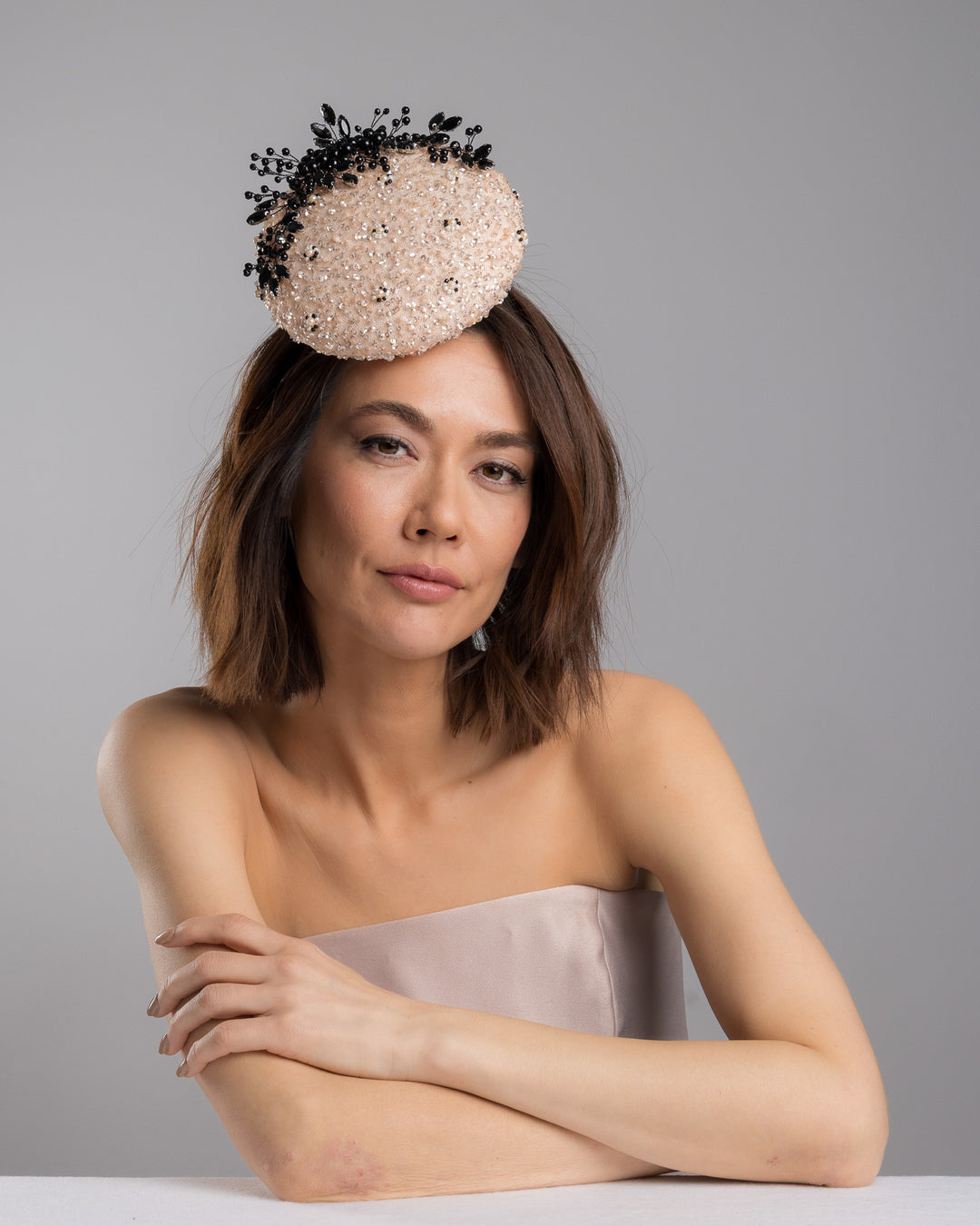 The female model is posing, wearing
Nuite, the blush sinamay fascinator with a rounded high-back silhouette, intricately embellished with clear crystals and pearl accents, and finished with a dramatic black beaded floral spray for elegant contrast. Designed and made by Merve Bayindir Millinery.