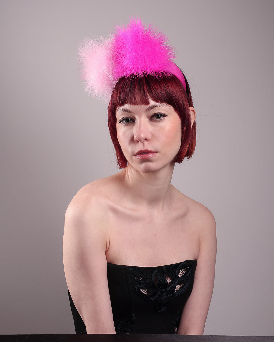 The model showcases the front left side of the NYX that is crafted on a hot pink satin headband. This playful fascinator features two pom-pom clouds of marabou feathers in graduated shades of candy pink and fuchsia. Designed by Merve Bayindir.