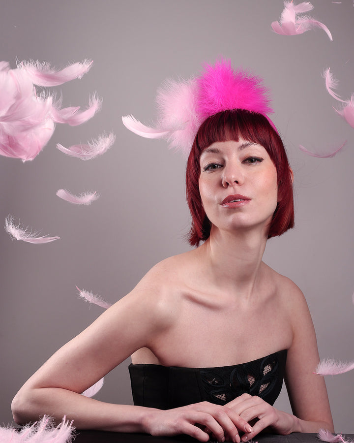 The model posing in the middle of flying feathers showcasing the front of the NYX that is crafted on a hot pink satin headband. This playful fascinator features two pom-pom clouds of marabou feathers in graduated shades of candy pink and fuchsia. Designed by Merve Bayindir.