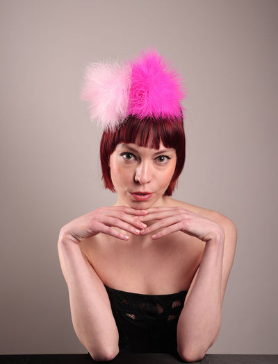 The model showcases the front of the NYX that is crafted on a hot pink satin headband. This playful fascinator features two pom-pom clouds of marabou feathers in graduated shades of candy pink and fuchsia. Designed by Merve Bayindir.