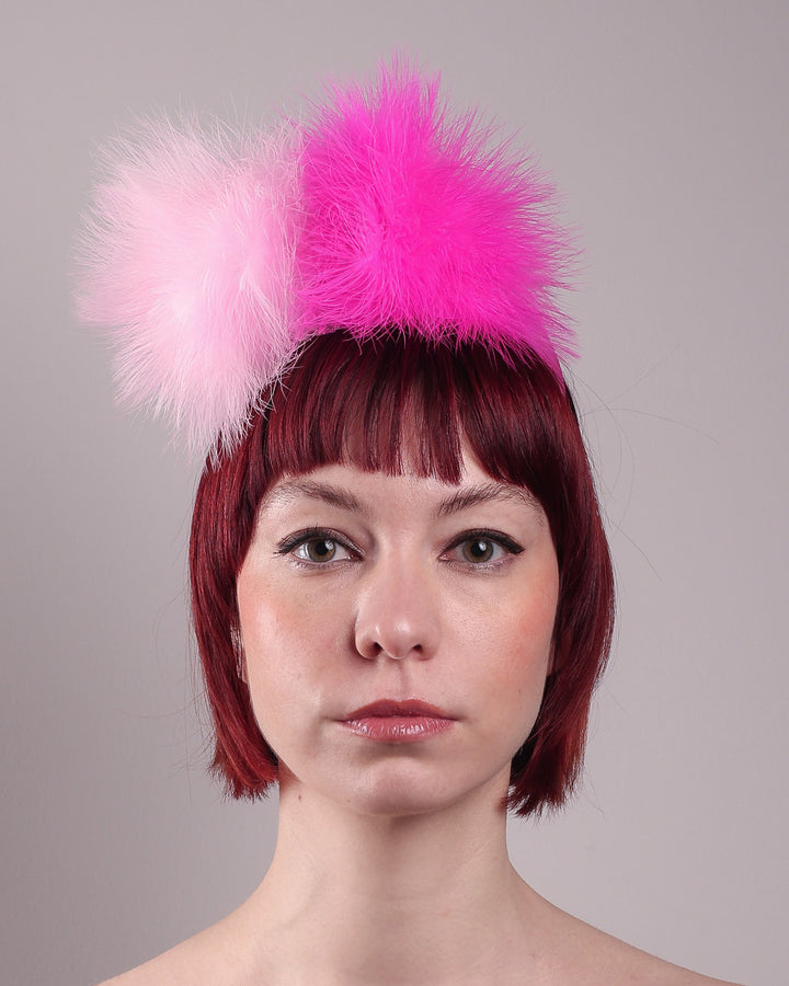 The model showcases the front close up of the NYX that is crafted on a hot pink satin headband. This playful fascinator features two pom-pom clouds of marabou feathers in graduated shades of candy pink and fuchsia. Designed by Merve Bayindir.