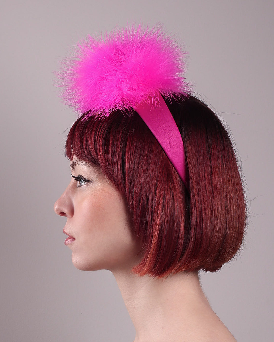 The model showcases the left side close up of the NYX that is crafted on a hot pink satin headband. This playful fascinator features two pom-pom clouds of marabou feathers in graduated shades of candy pink and fuchsia. Designed by Merve Bayindir.