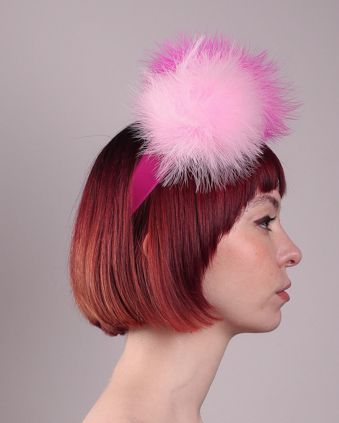 The model showcases the left side close up of the NYX that is crafted on a hot pink satin headband. This playful fascinator features two pom-pom clouds of marabou feathers in graduated shades of candy pink and fuchsia. Designed by Merve Bayindir.