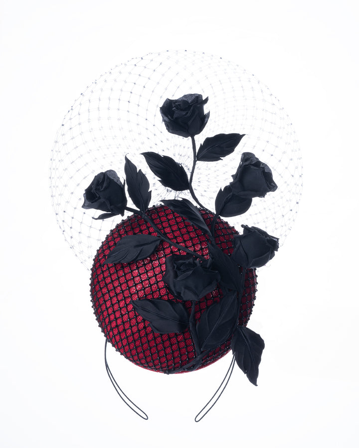 A bold fascinator composed of a structured circular base in deep red, overlaid with black netting that creates a texture. From the base an  arrangement of matte black roses and elongated leaves, climbing upward in a graceful, vine-like composition.Behind the florals, a sheer circular veil of fine netting forms a delicate halo. Designed and made by Merve Bayindir Millinery.
