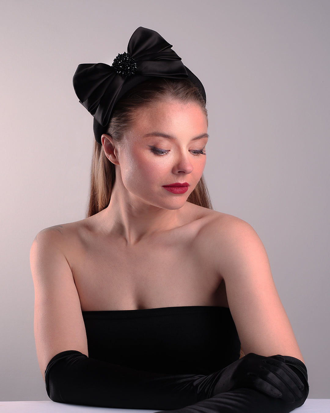 The model is showcasing the right front angle of a high black headband covered with lycra fabric decorated with two layers of sateen bow on the right side. The bow is embellished with black crystal beads in the center.
