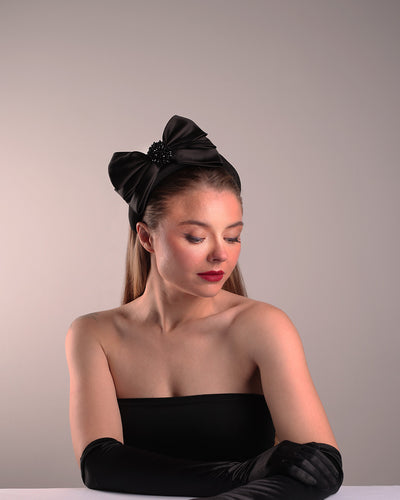 The model is showcasing the right front angle of a high black headband covered with lycra fabric decorated with two layers of sateen bow on the right side. The bow is embellished with black crystal beads in the center.