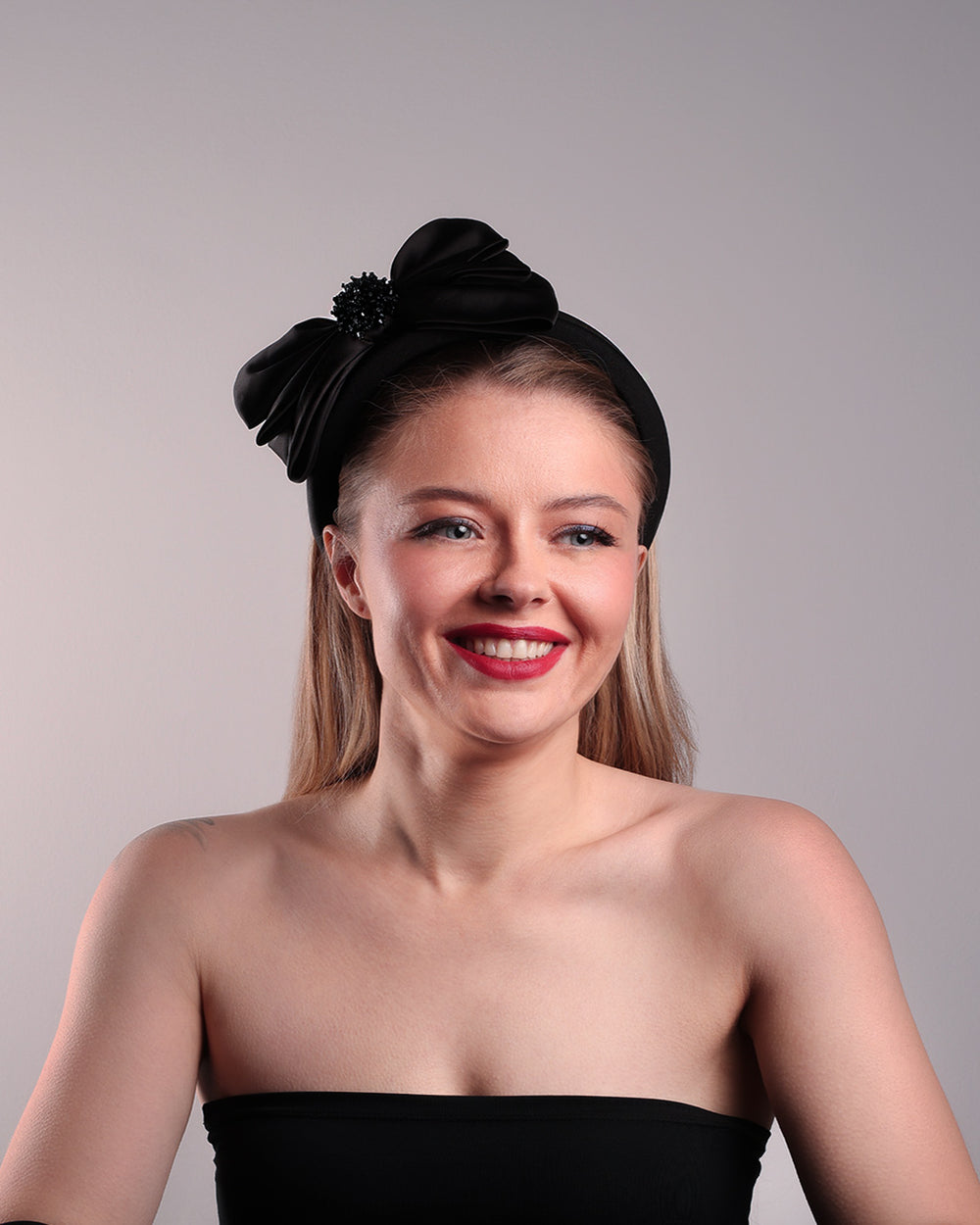 The model is showcasing the front of a high black headband covered with lycra fabric decorated with two layers of sateen bow on the right side. The bow is embellished with black crystal beads in the center.
