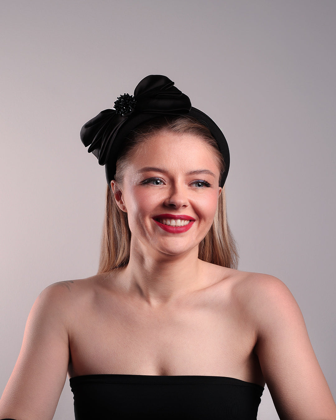 The model is showcasing the front of a high black headband covered with lycra fabric decorated with two layers of sateen bow on the right side. The bow is embellished with black crystal beads in the center.
