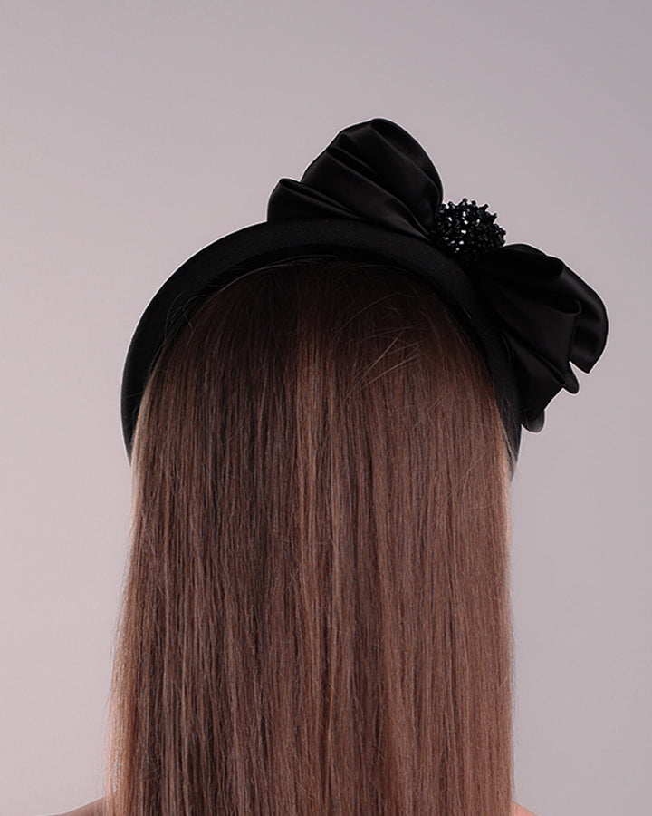 The model is showcasing the left side of a high black headband covered with lycra fabric decorated with two layers of sateen bow on the right side. The bow is embellished with black crystal beads in the center.