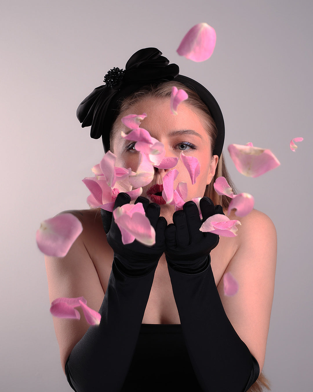 The model is showcasing the right side of a high black headband covered with lycra fabric decorated with two layers of sateen bow on the right side while blowing rose petals. An editorial picture.