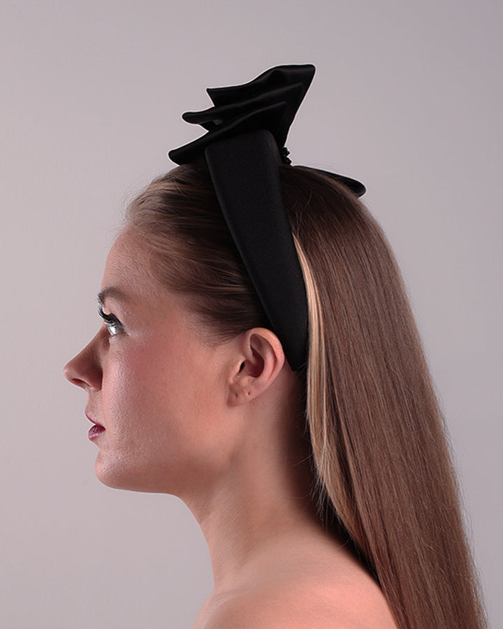 The model is showcasing the left side of a high black headband covered with lycra fabric decorated with two layers of sateen bow on the right side. The bow is embellished with black crystal beads in the center.