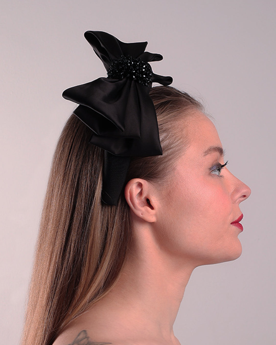 The model is showcasing the right side of a high black headband covered with lycra fabric decorated with two layers of sateen bow on the right side. The bow is embellished with black crystal beads in the center.