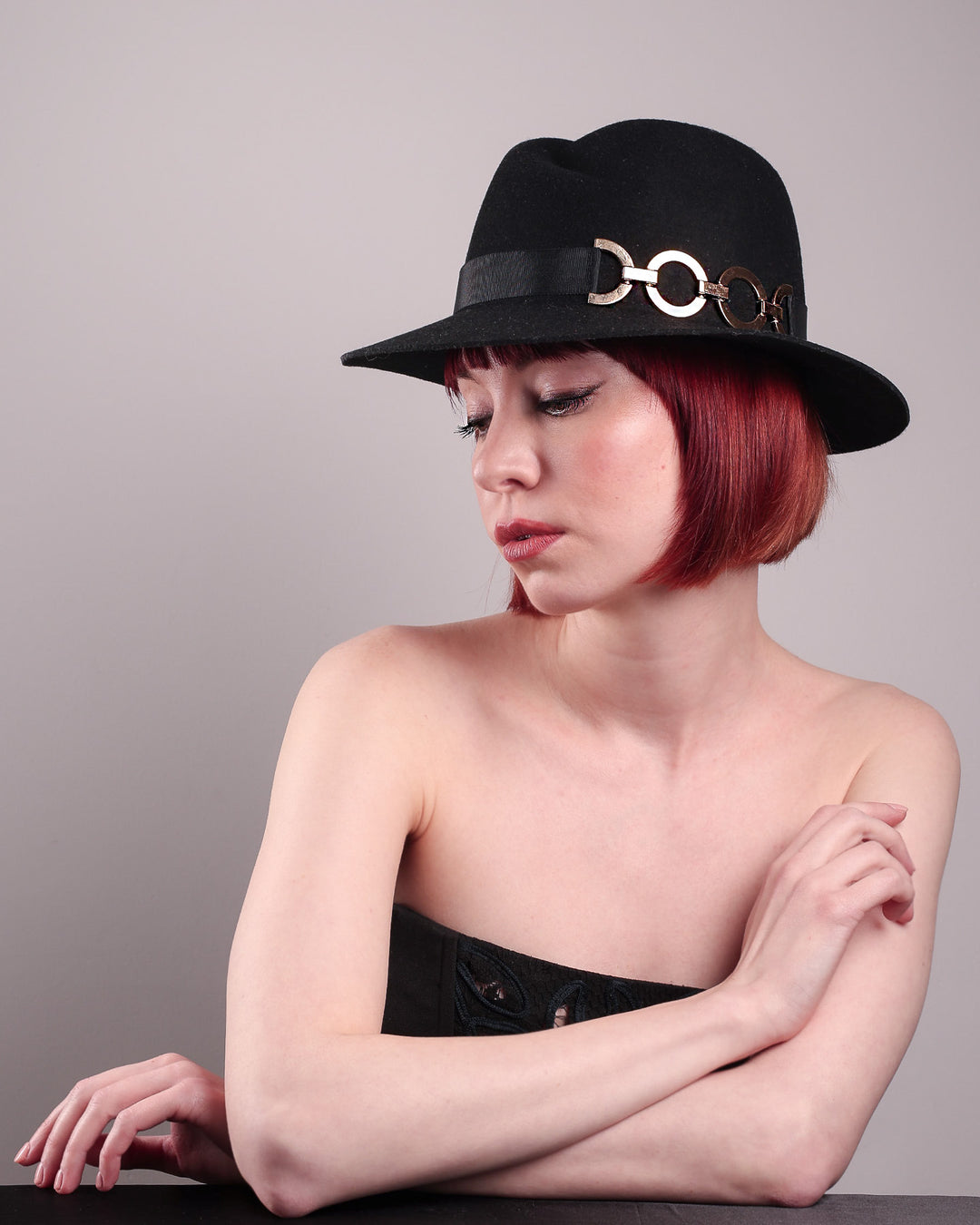 The model showcases the left front side of the Nocturne Fedora hat, crafted from rich black wool felt. This structured silhouette is elevated by a gold coloured, bold metallic chain detail in warm gold on the left side, linked seamlessly into a sleek black grosgrain band.