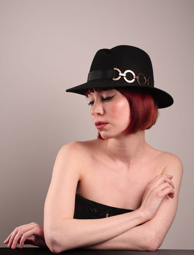 The model showcases the left front side of the Nocturne Fedora hat, crafted from rich black wool felt. This structured silhouette is elevated by a gold coloured, bold metallic chain detail in warm gold on the left side, linked seamlessly into a sleek black grosgrain band.