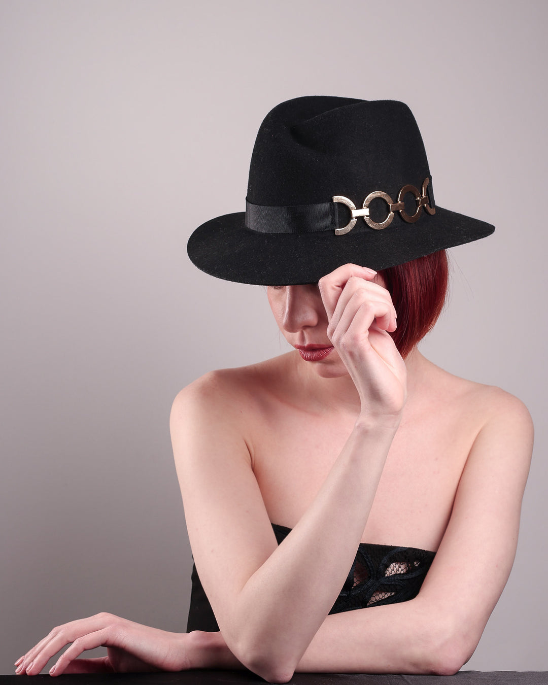 The model showcases the left front from the above angle of the Nocturne Fedora hat, crafted from rich black wool felt. This structured silhouette is elevated by a gold coloured, bold metallic chain detail in warm gold on the left side, linked seamlessly into a sleek black grosgrain band.