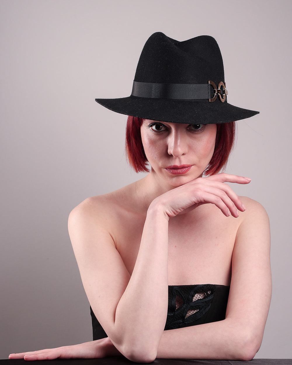 The model showcases the front of the Nocturne Fedora hat, crafted from rich black wool felt. This structured silhouette is elevated by a gold coloured, bold metallic chain detail in warm gold on the left side, linked seamlessly into a sleek black grosgrain band.